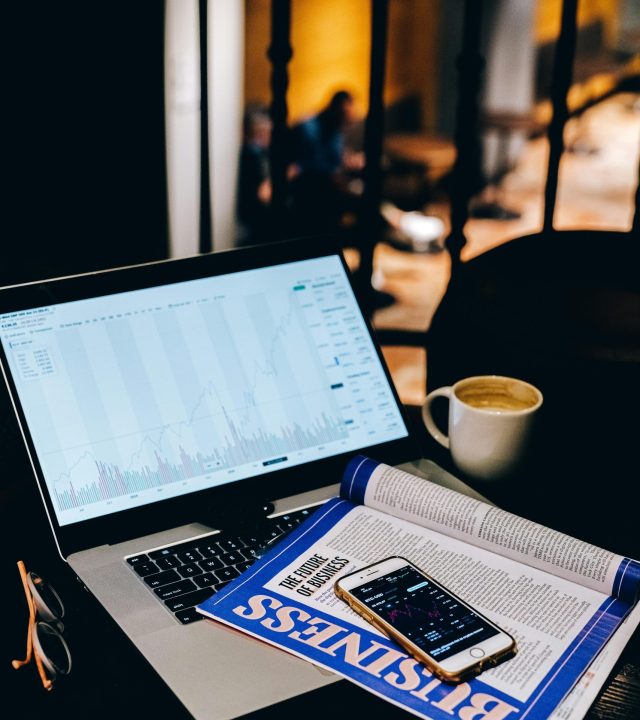 A laptop showing financial charts beside a business magazine and coffee cup in a cafe setting.